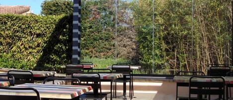 An outdoor patio with tables and chairs, surrounded by dense foliage and a clear sky.