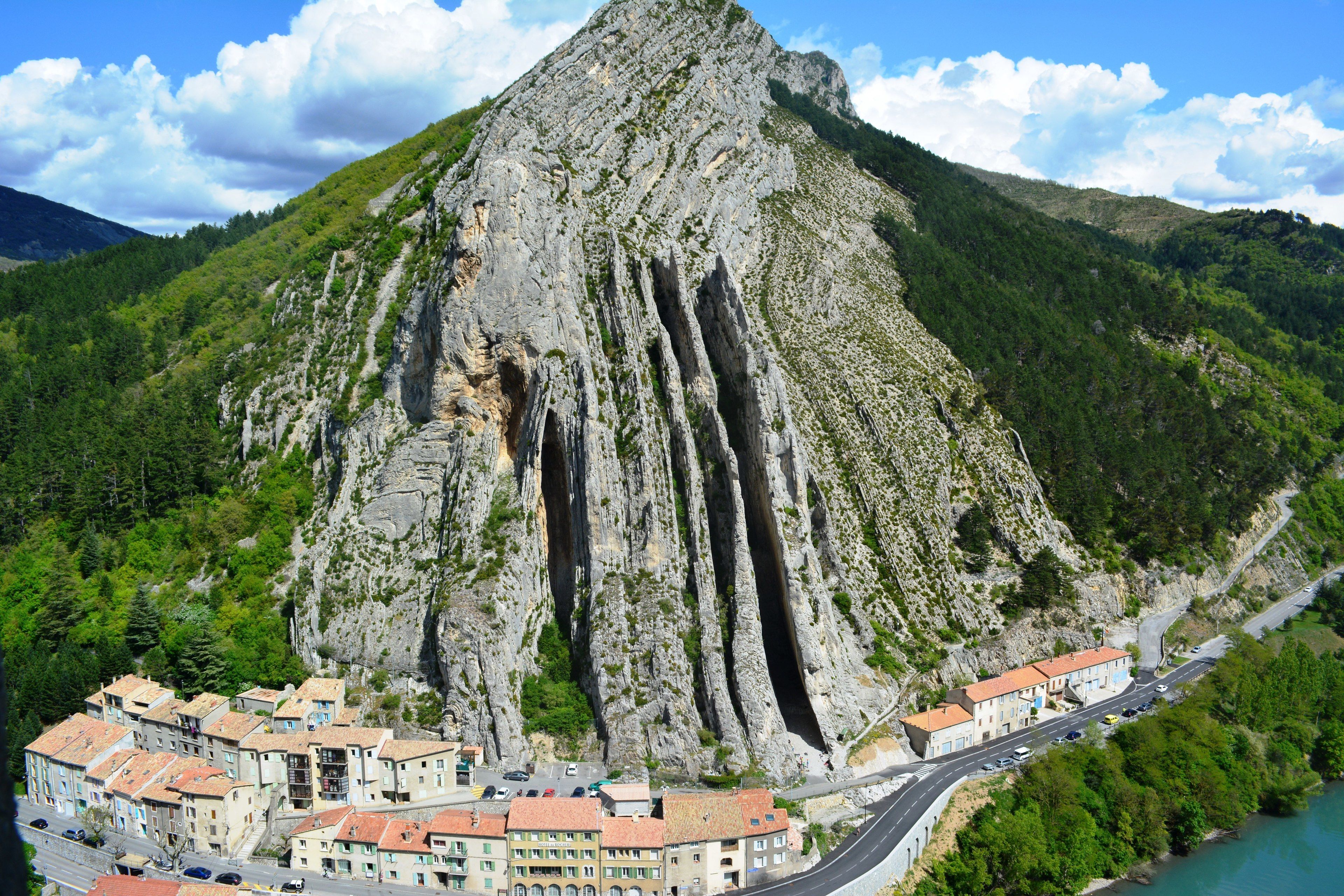 A mountainous landscape with a prominent rocky peak, a winding road, and a cluster of buildings with red-tiled roofs.