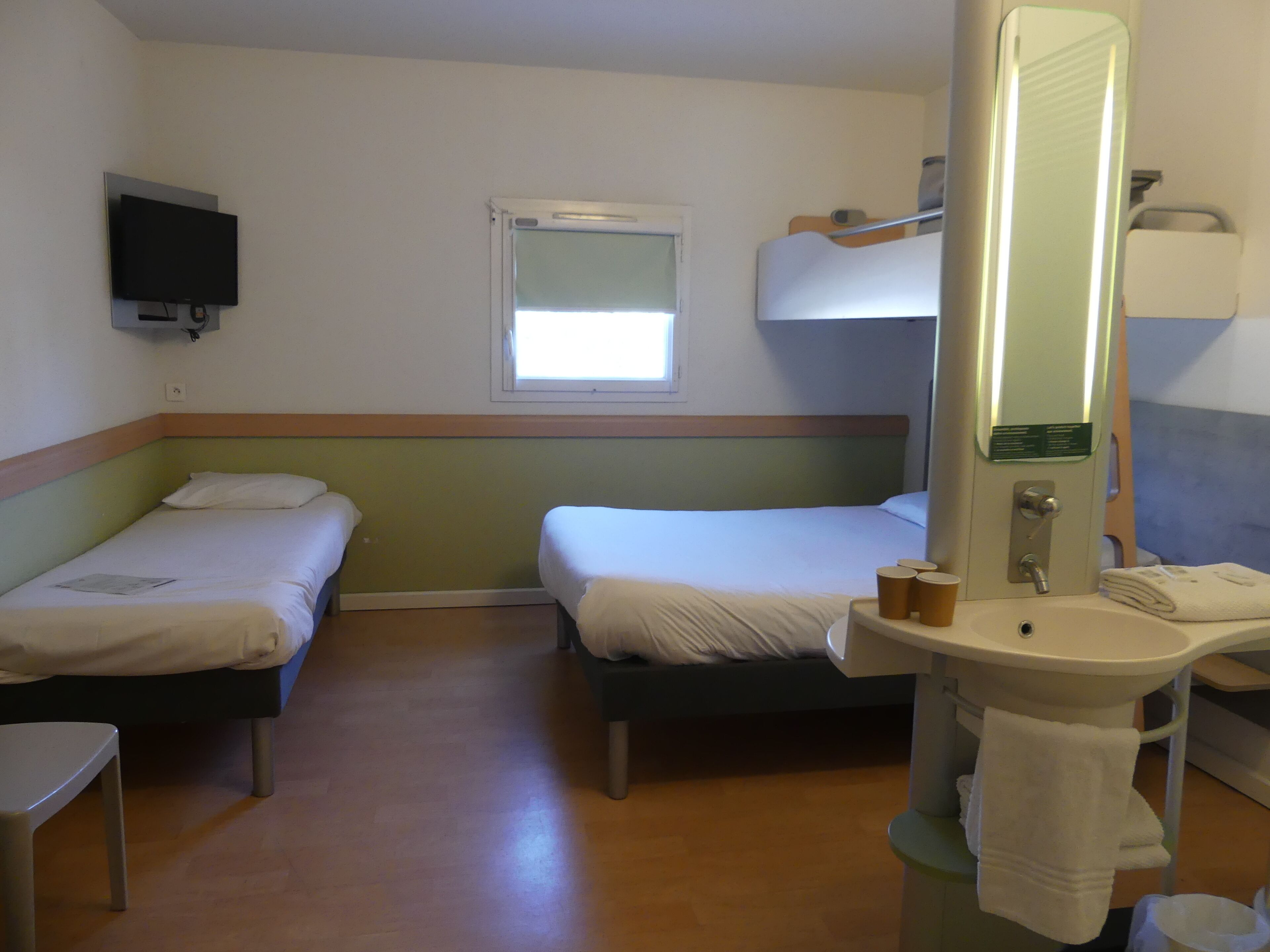 A hotel room with two single beds, a small table, a TV mounted on the wall, a window, and a bathroom with a sink and mirror.