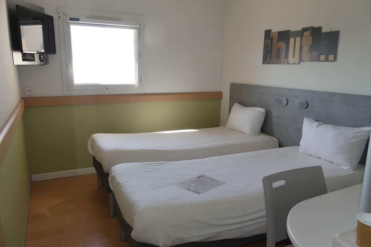 A hotel room with two beds, a small table, a chair, a window, and a wall-mounted light fixture.