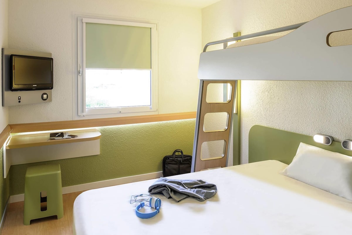 A hotel room with a bunk bed, a desk with a chair, a television, and a window.