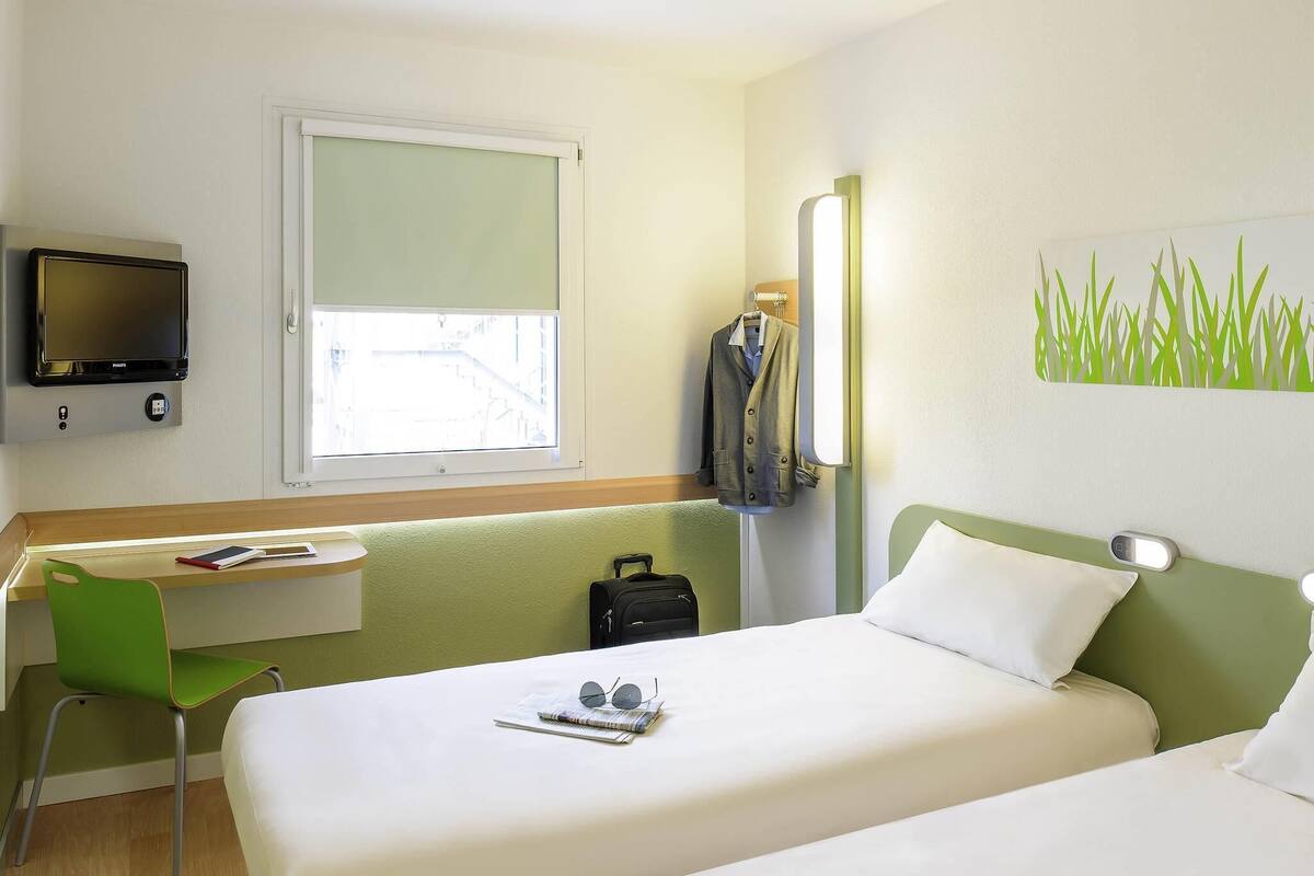 A hotel room with two beds, a desk, a chair, a TV, and a window with a view of buildings.