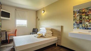 A neatly arranged hotel room with a bed, a desk, a chair, a wall-mounted TV, and a poster wall.