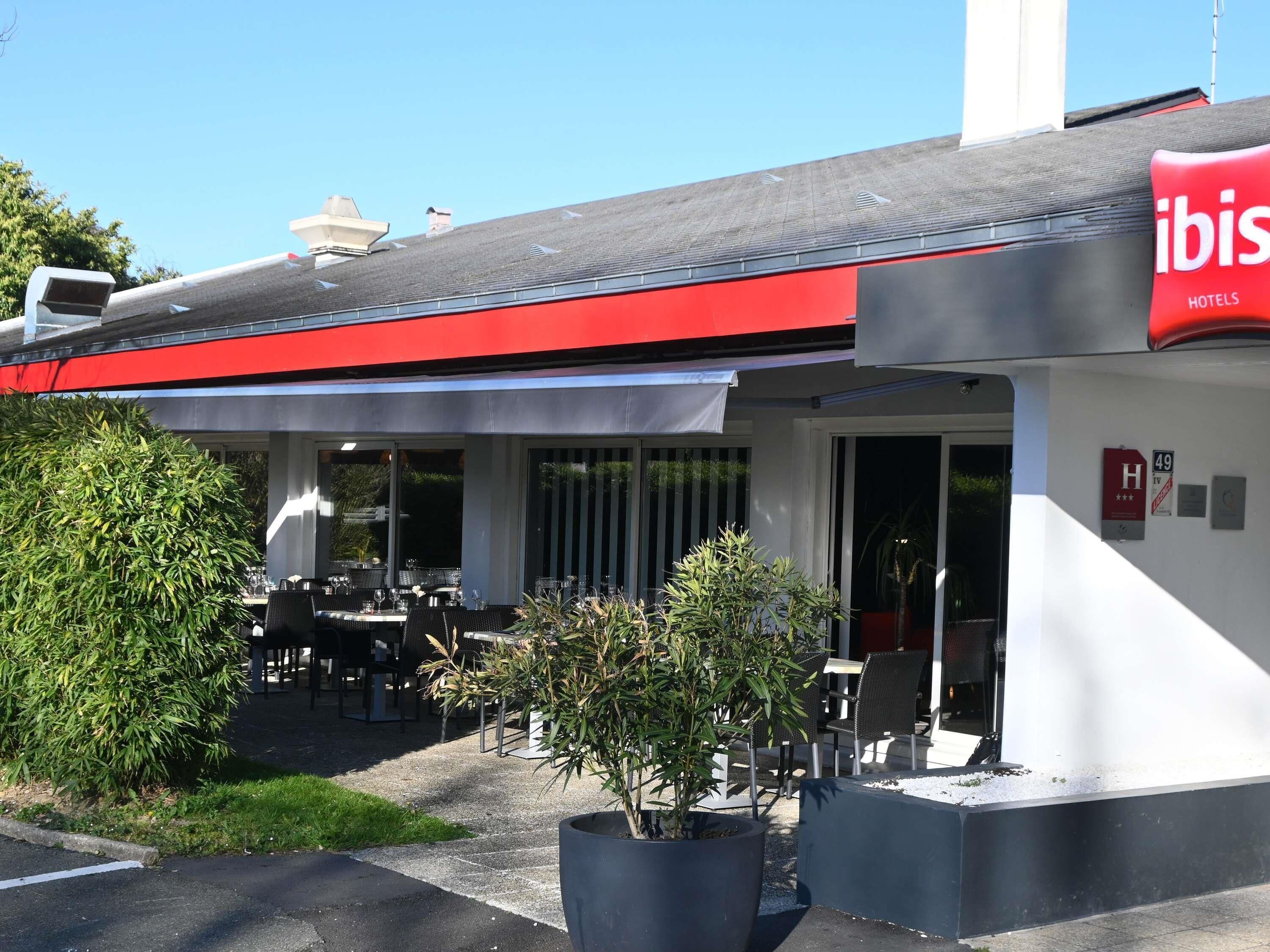 Un área de estar al aire libre con mesas y sillas, una planta en maceta, y un edificio con toldo rojo y un letrero que dice 'ibis HOTELS'.