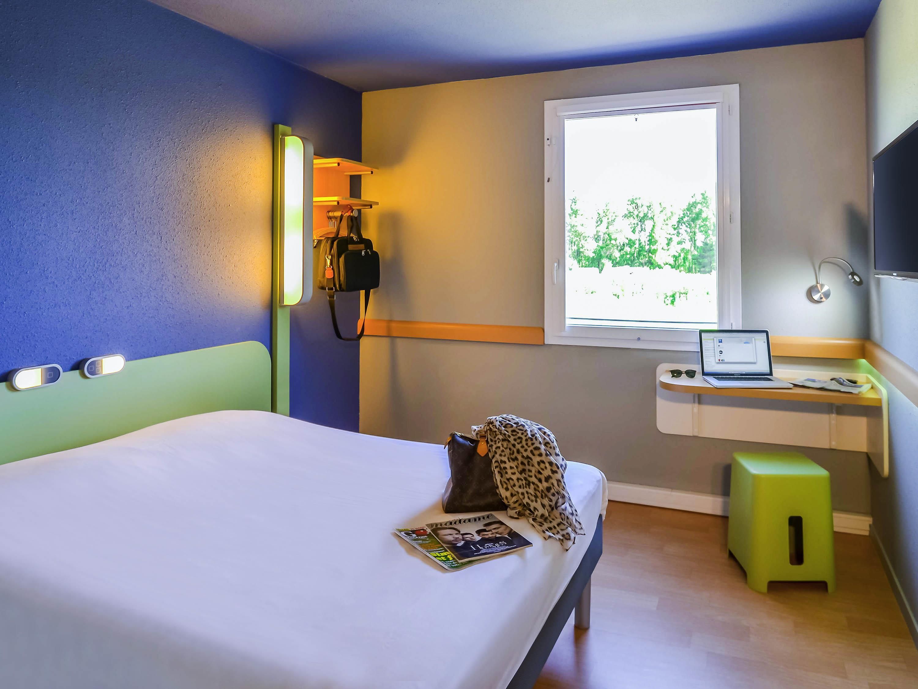 A hotel room with a bed, a desk with a laptop, a window with a view, and a bedside lamp.