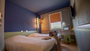 A bedroom with two beds, a desk, a chair, a television, and a window with a view of greenery. - ibis budget Saint Martin de Crau Porte de Camargue (Saint-Martin-de-Crau)