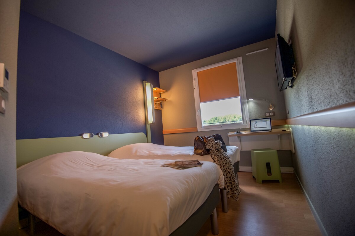 A bedroom with two beds, a desk, a chair, a television, and a window with a view of greenery.