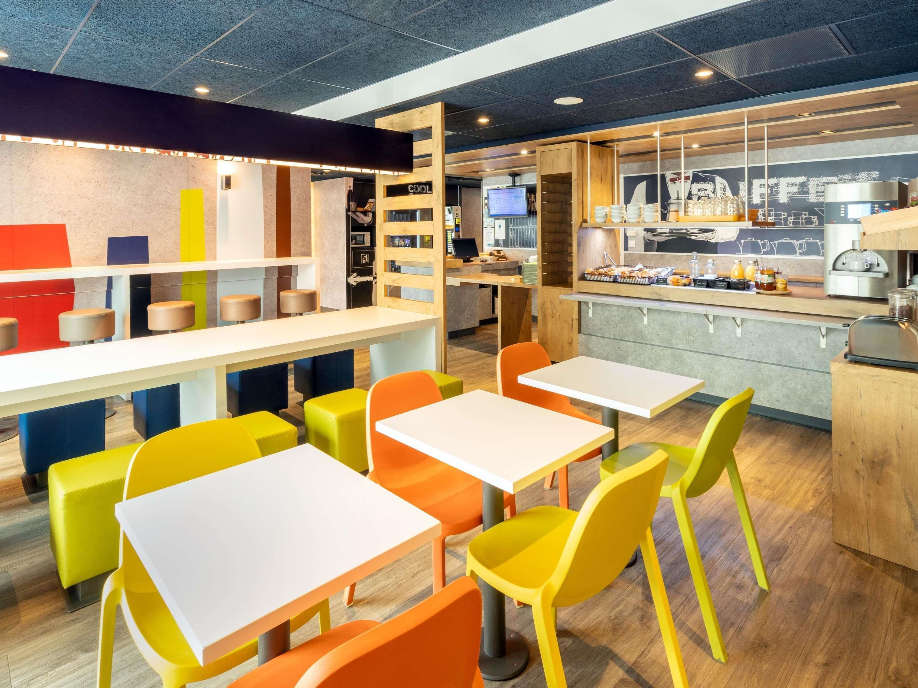 Interior moderno de una cafetería con sillas y mesas de colores, una barra con máquina de café y un cartel con el menú.