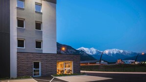 Ein modernes Gebäude mit sichtbarer Essecke durch die Fenster, einem Parkplatz und einer Bergkulisse.