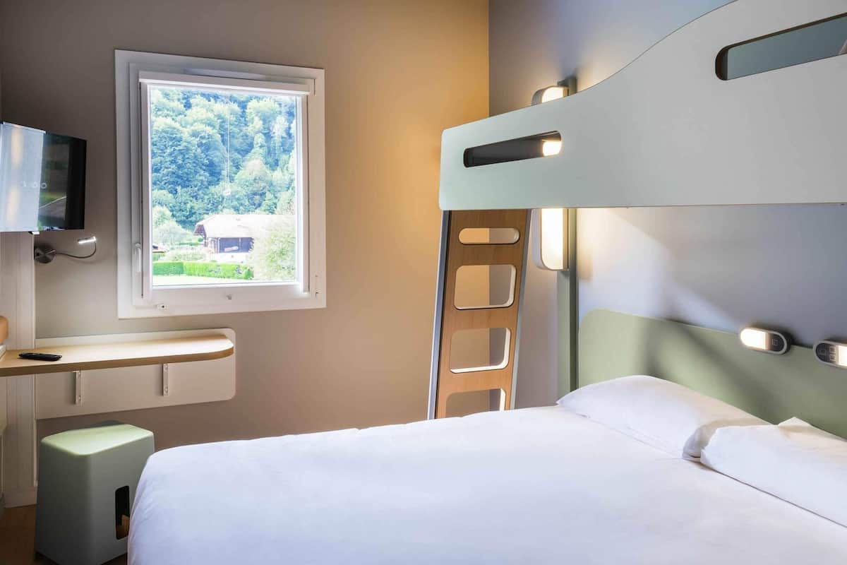 Une chambre d’hôtel moderne équipée d’un lit superposé, d’un bureau avec une chaise, d’une télévision et d’une fenêtre donnant sur un paysage verdoyant.