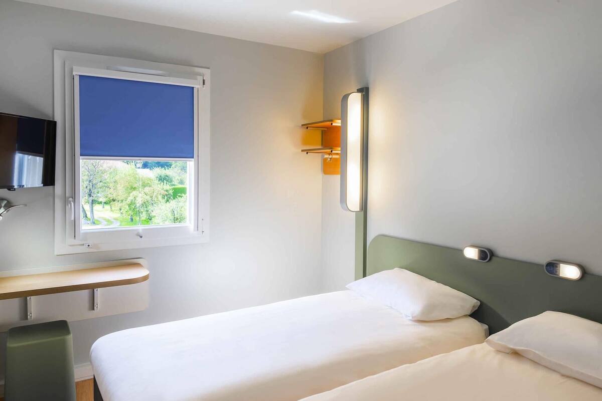 Une chambre d’hôtel comprenant un lit, un bureau, une chaise, une fenêtre donnant sur un espace verdoyant et un luminaire fixé au mur.
