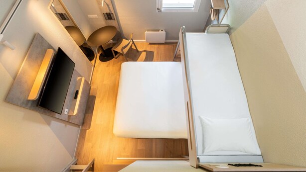 Une chambre d’hôtel de taille réduite, comprenant un lit, un bureau avec un ordinateur, une chaise, une petite table, une fenêtre et un luminaire au plafond.