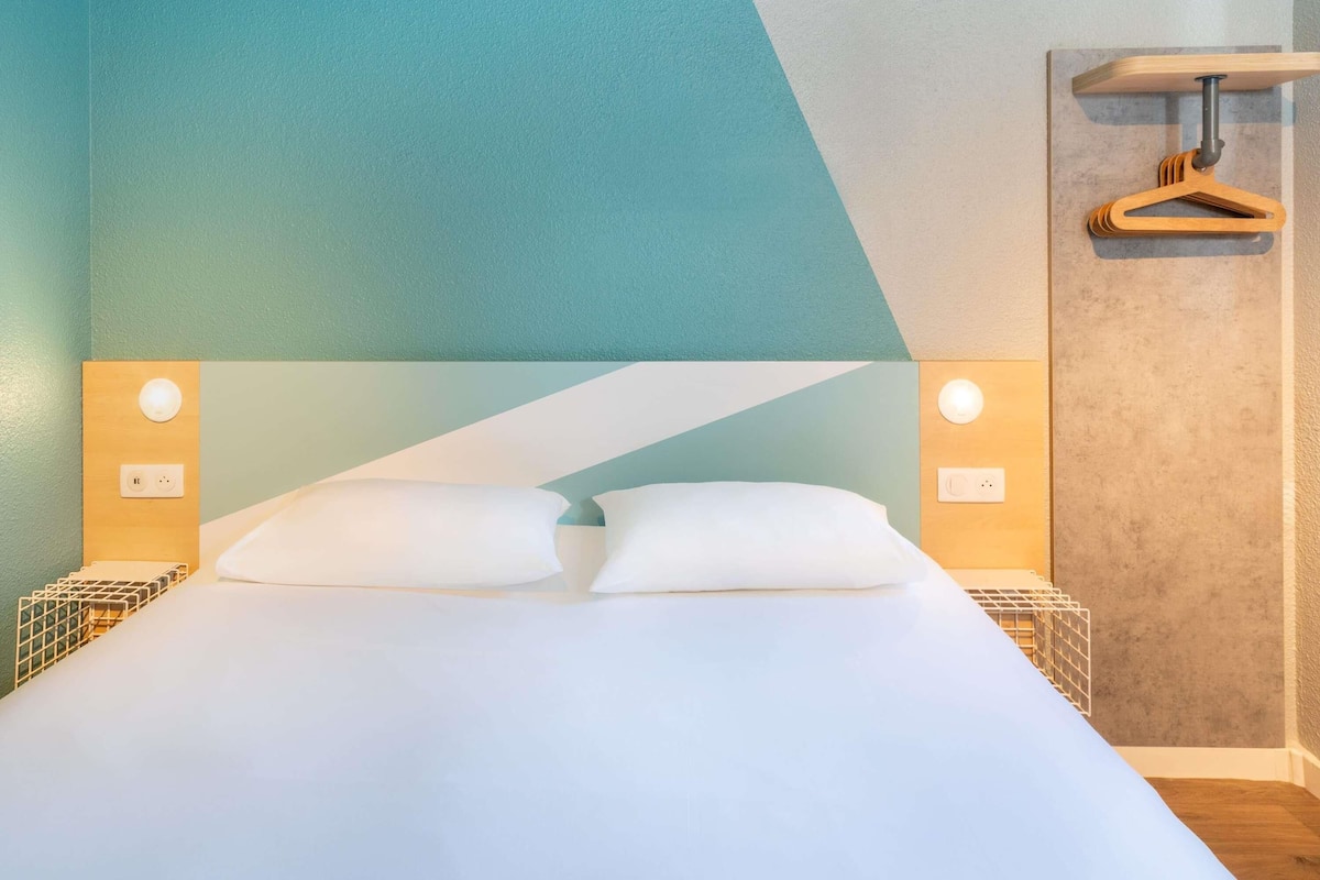 Un lit avec du linge de lit blanc, deux tables de chevet avec des lampes, un porte-manteau en bois et un motif géométrique sur le mur.