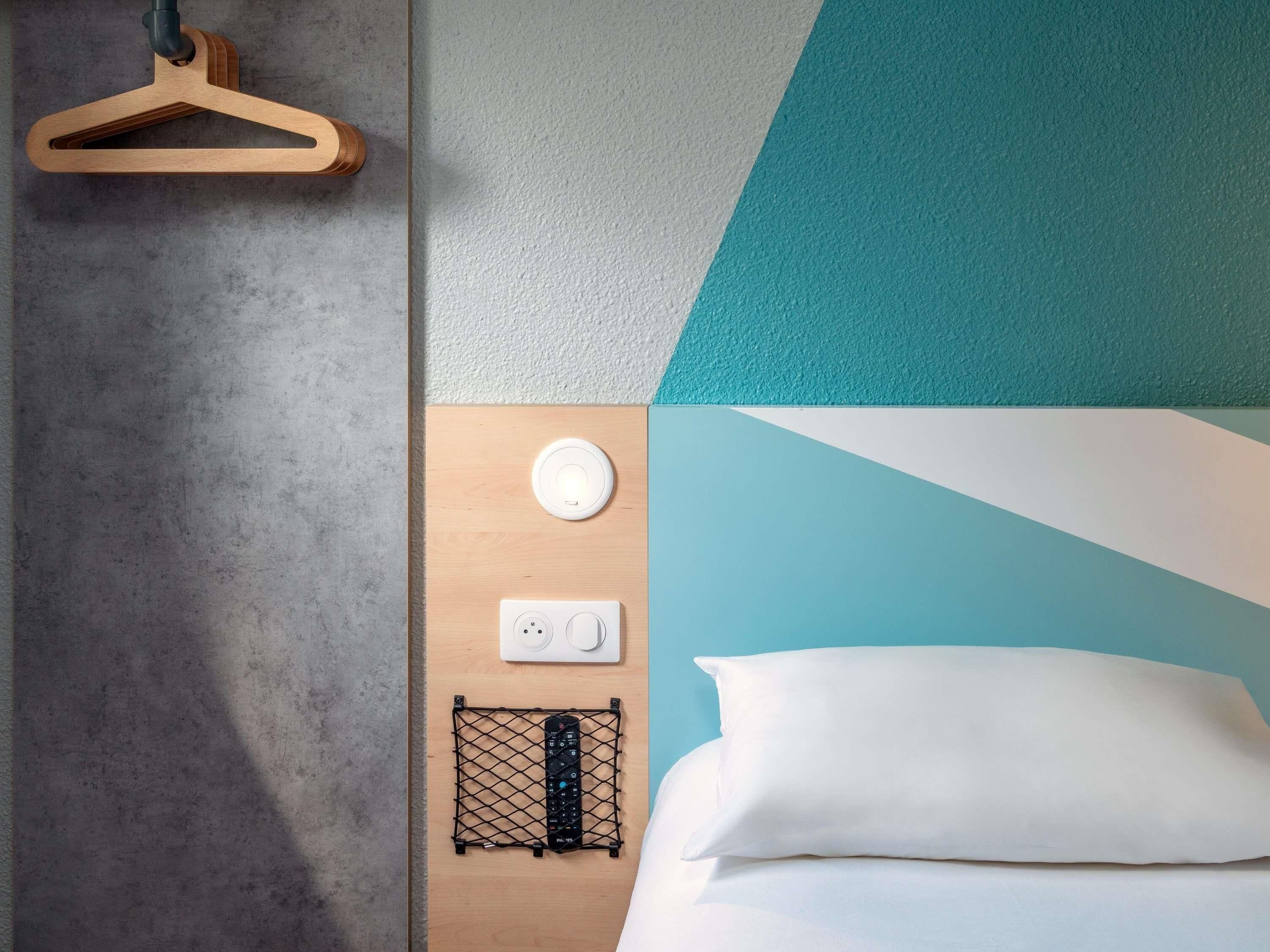 A neatly arranged hotel room with a wooden bedside table, a power outlet, a remote control on a wire shelf, a white pillow, and a hanging coat hook.