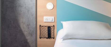 A neatly arranged hotel room with a wooden bedside table, a power outlet, a remote control on a wire shelf, a white pillow, and a hanging coat hook.
