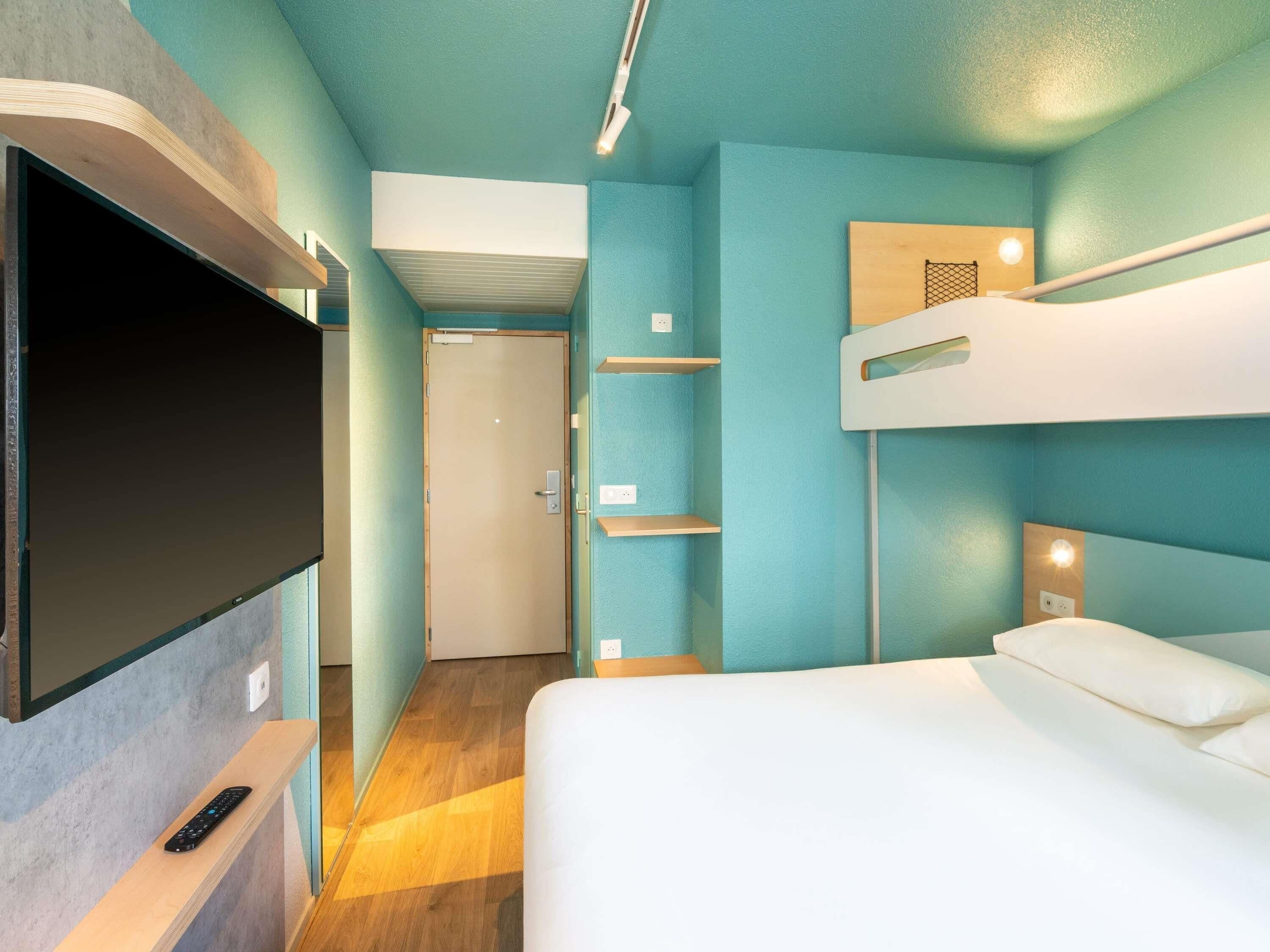 Quarto de hotel moderno com televisão de tela plana, beliches, uma prateleira de madeira e uma porta que dá para outro cômodo.