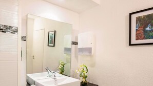 A bathroom with a white sink, two faucets, a mirror, a framed picture, a wall-mounted light, and a small vase with flowers. - ibis budget Antibes Sophia Antipolis (Vallauris)
