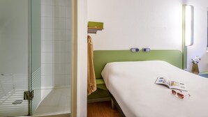 A hotel room with a glass shower enclosure, a bed with a green headboard, a bedside table with a book and sunglasses, and a wall-mounted light fixture. - ibis budget Antibes Sophia Antipolis (Vallauris)