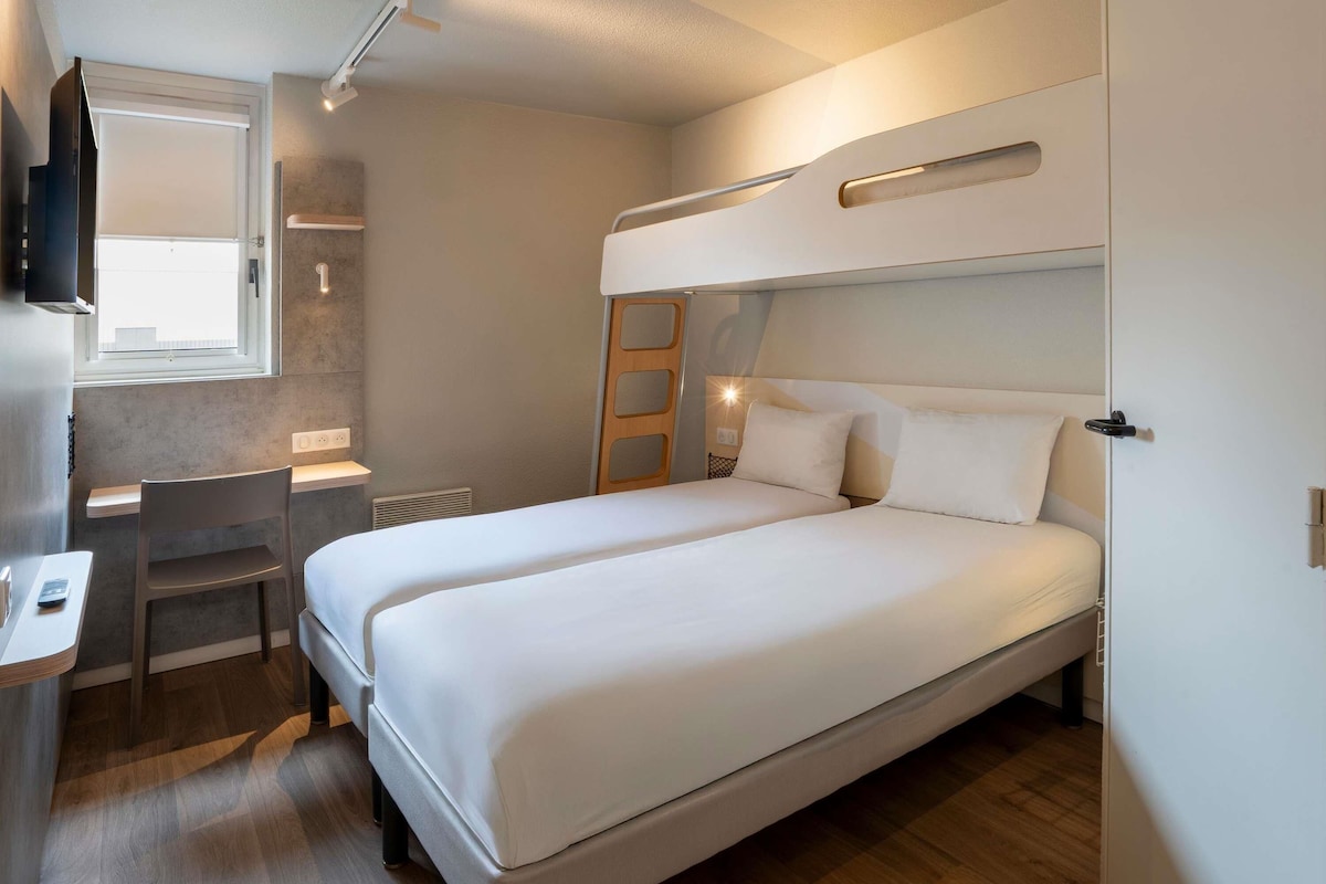 A compact room with a bunk bed, a desk with a chair, a small window, and a wall-mounted TV.
