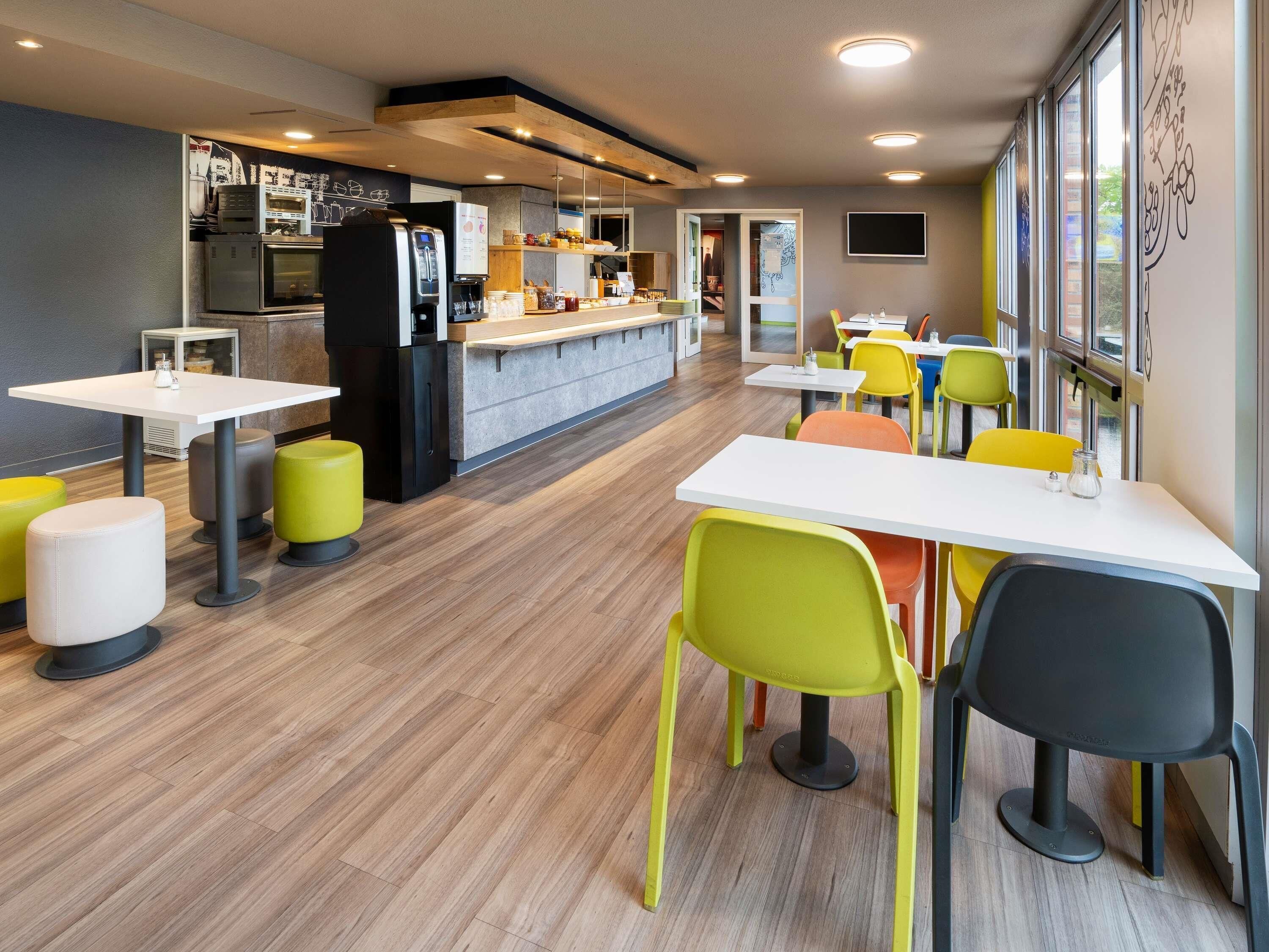 A modern dining area with tables and chairs, a kitchen in the background, and a view through the window.