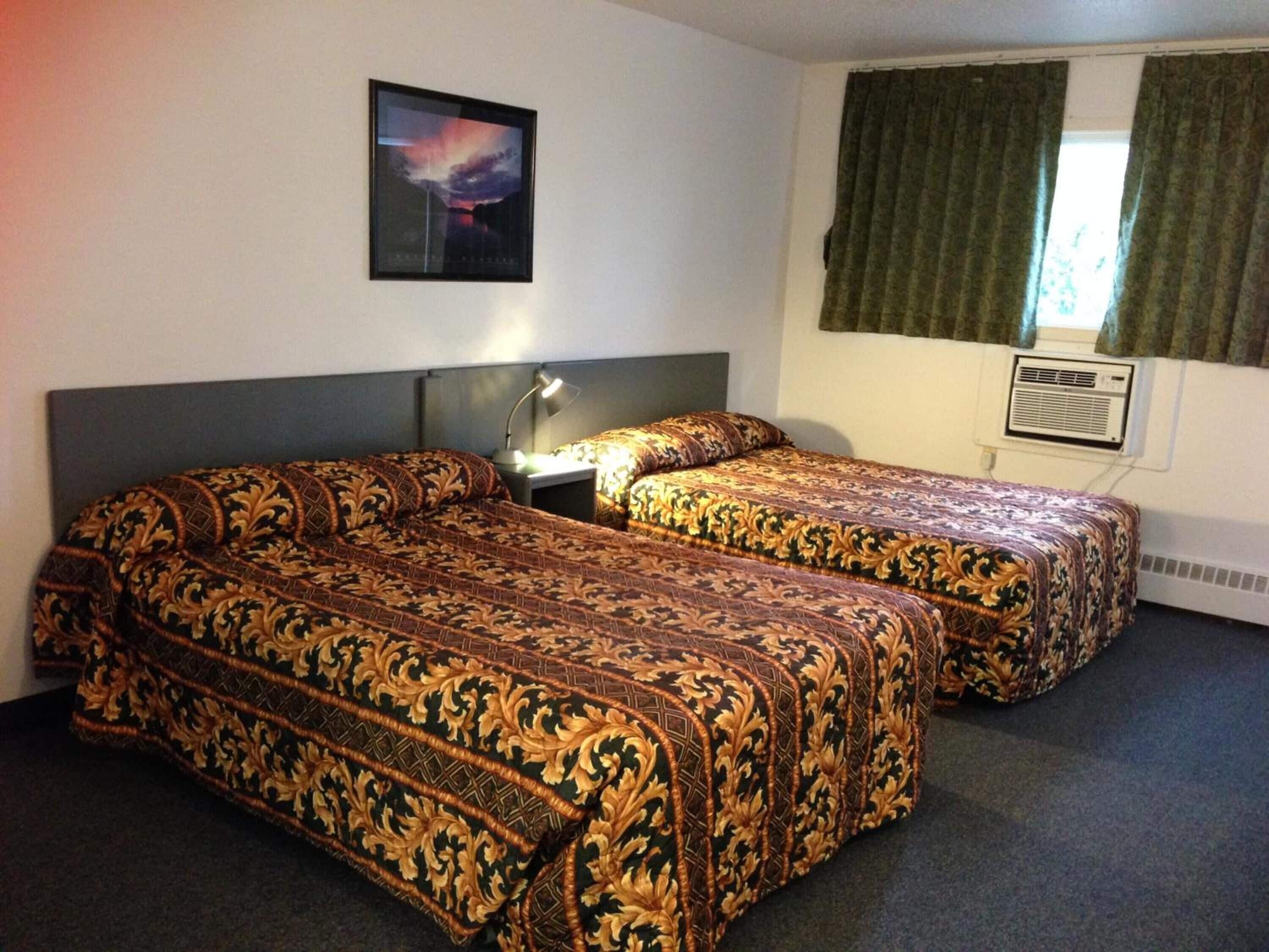 A hotel room with two beds, a nightstand, a lamp, a picture on the wall, and an air conditioning unit.