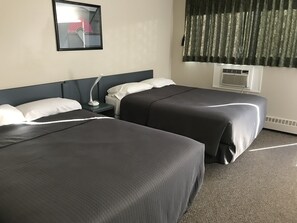 A hotel room with two beds, a nightstand, a lamp, a mirror, a window with curtains, and an air conditioning unit.