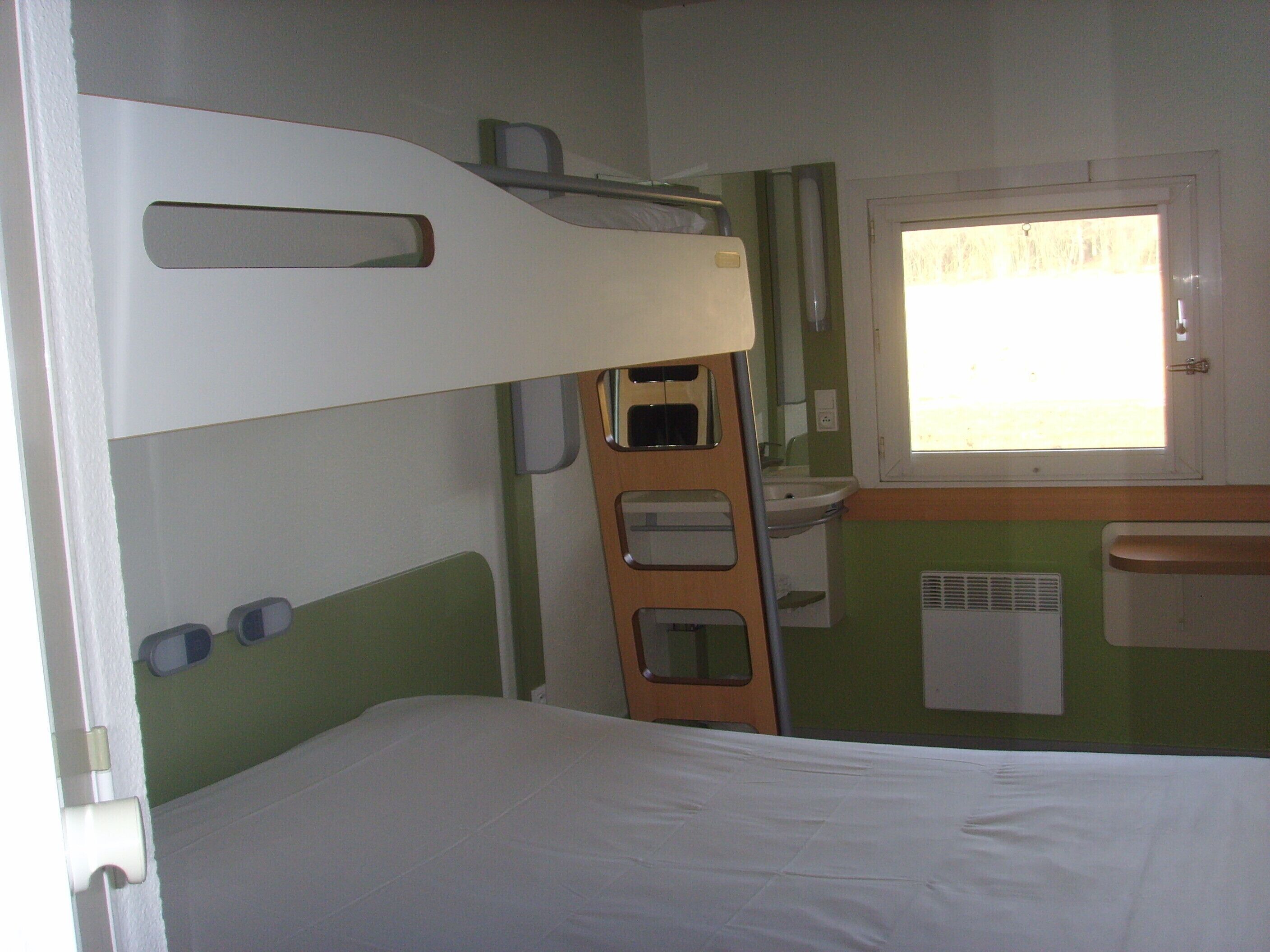 Una cama individual superpuesta con escalera, una ventana con cortinas, un lavabo y un radiador.