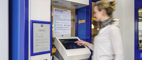 Punto de registro con un quiosco digital, un cartel que dice 'CHECK-IN POINT' y '¡BIENVENIDOS! ibis Budget!', y una ranura para 'TICKET'.
