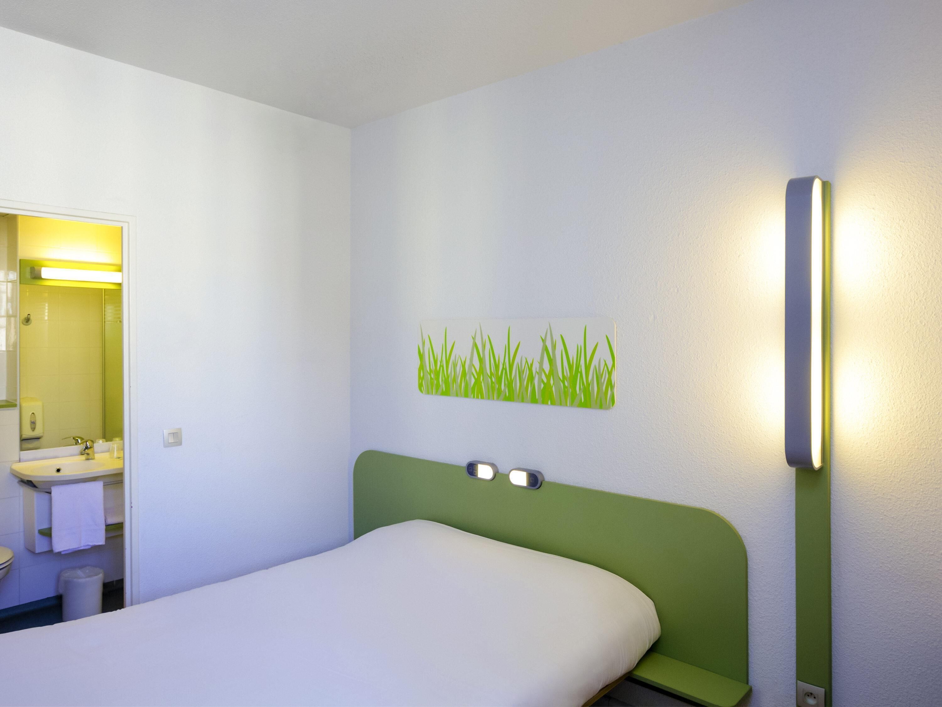 A hotel room with a bed, a green headboard, a wall-mounted light, and a bathroom visible through an open door.
