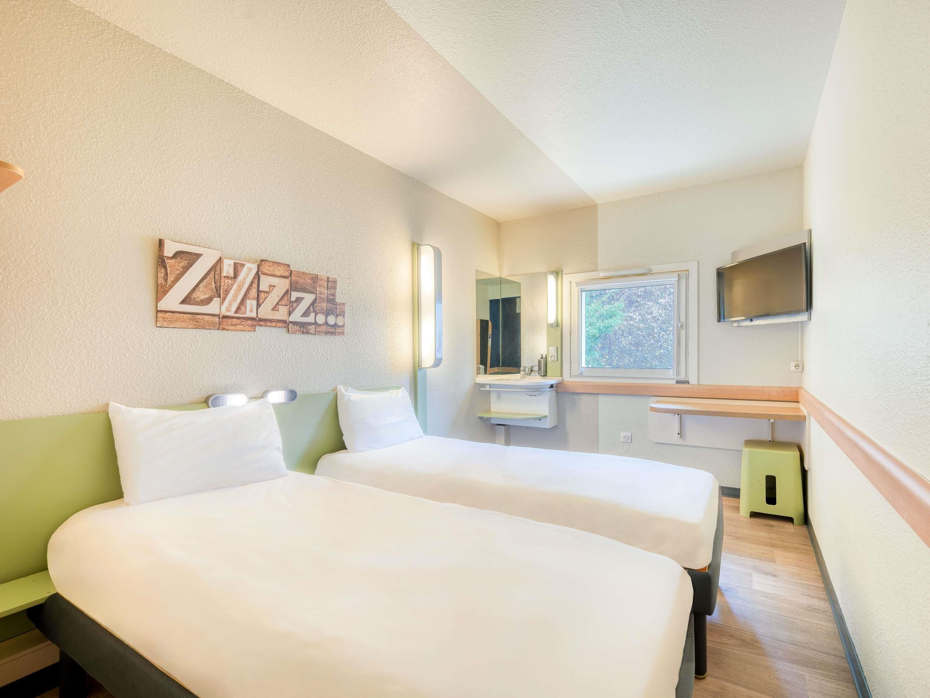 A hotel room with two beds, a small bathroom, a TV, and a window with a view of trees.