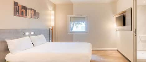 A modern hotel room with a bed, bedside table, lamp, TV, and a window.