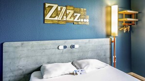 A hotel room with a bed, a headboard, a wall-mounted light, and a wall decoration. - ibis budget Versailles Trappes (Trappes)