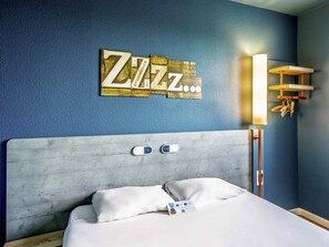 A hotel room with a bed, a headboard, a wall-mounted light, and a wall decoration. - ibis budget Versailles Trappes (Trappes)