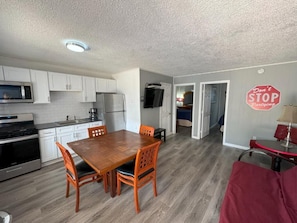Cocina compacta con armarios blancos, una mesa de comedor con sillas, un sofá rojo y un televisor montado en la pared.