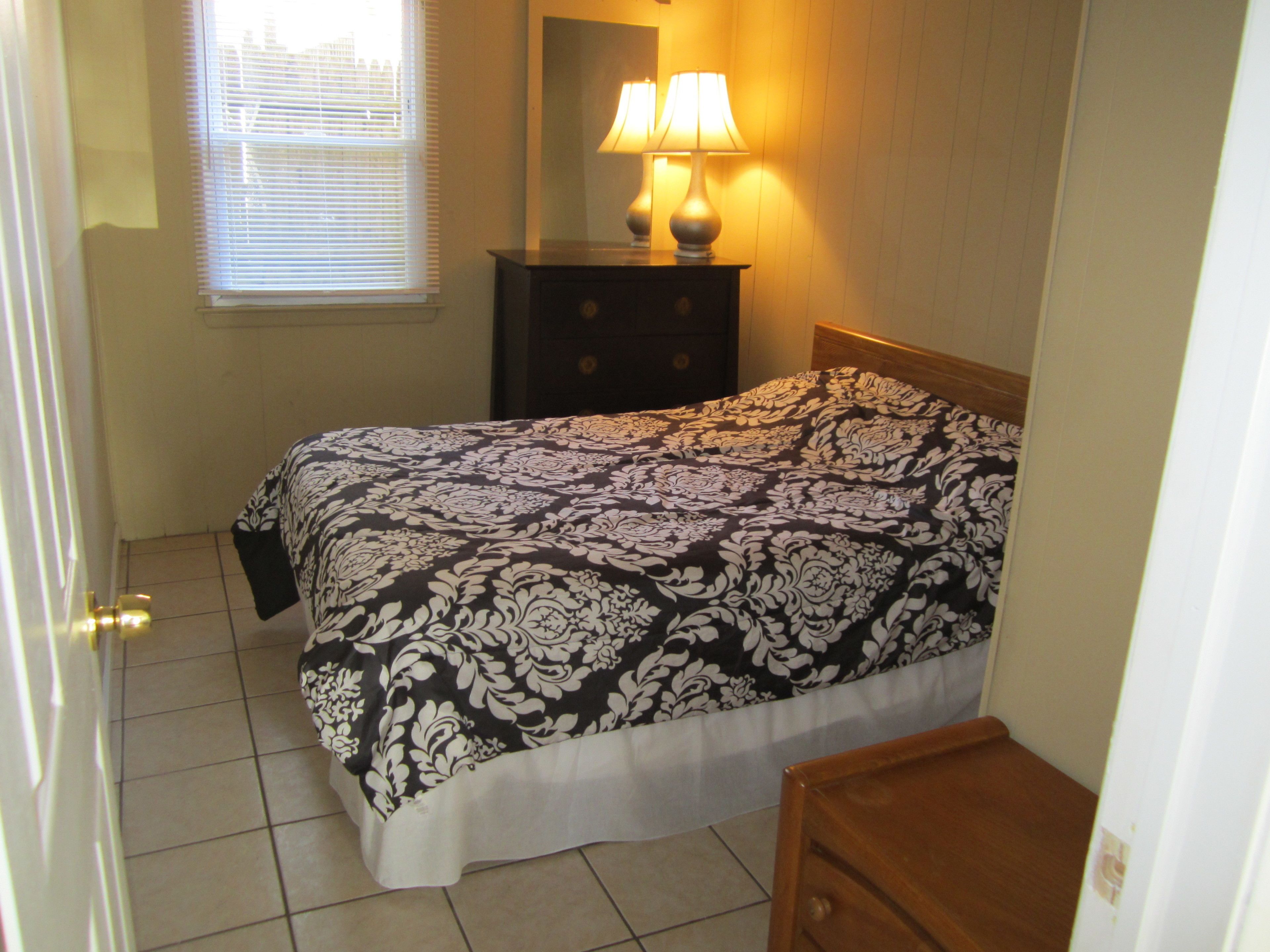A bedroom with a bed, a nightstand with a lamp, a window with blinds, and a door.