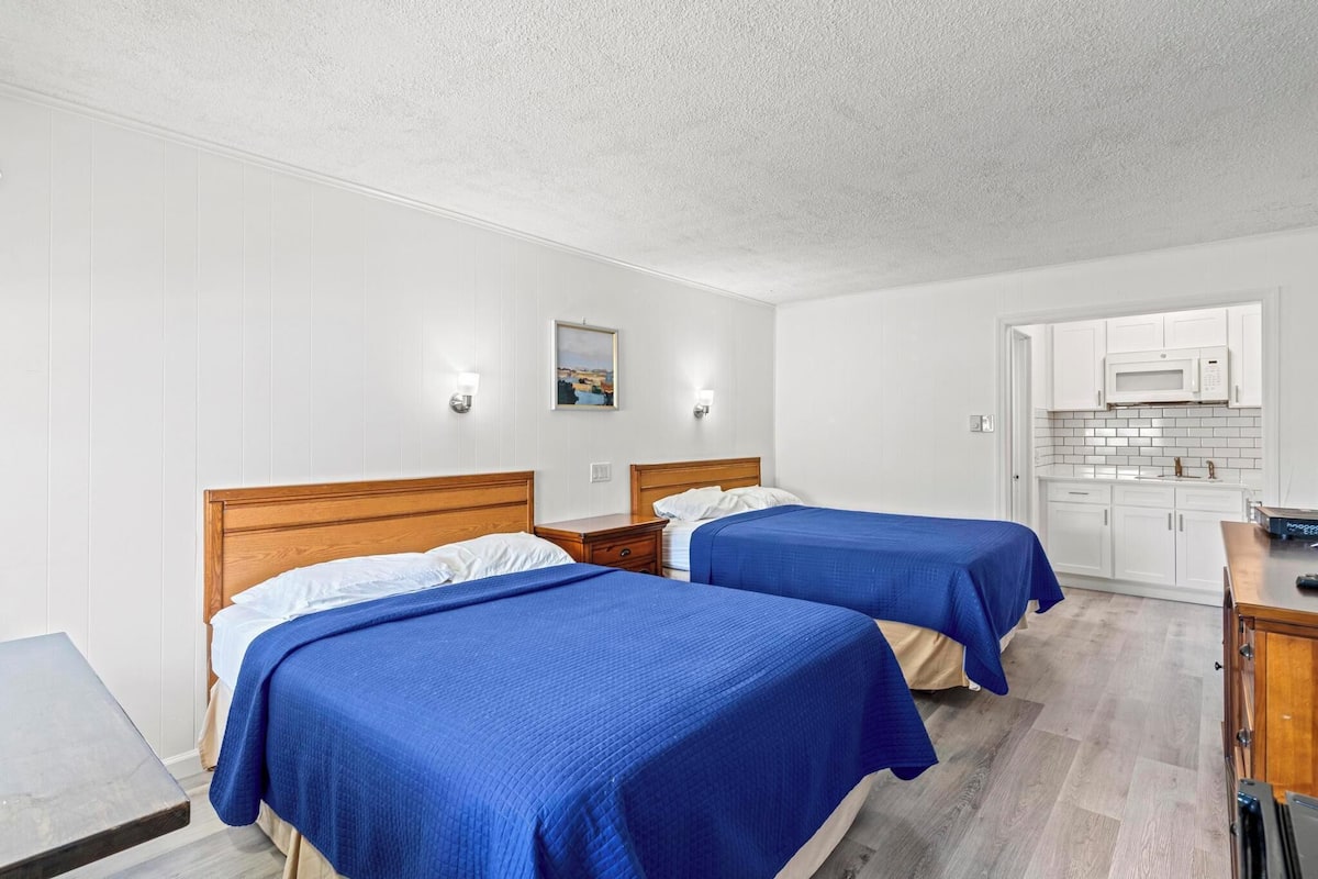 A hotel room with two beds, a wooden headboard, a small table, a microwave, and a tiled backsplash in the kitchenette.