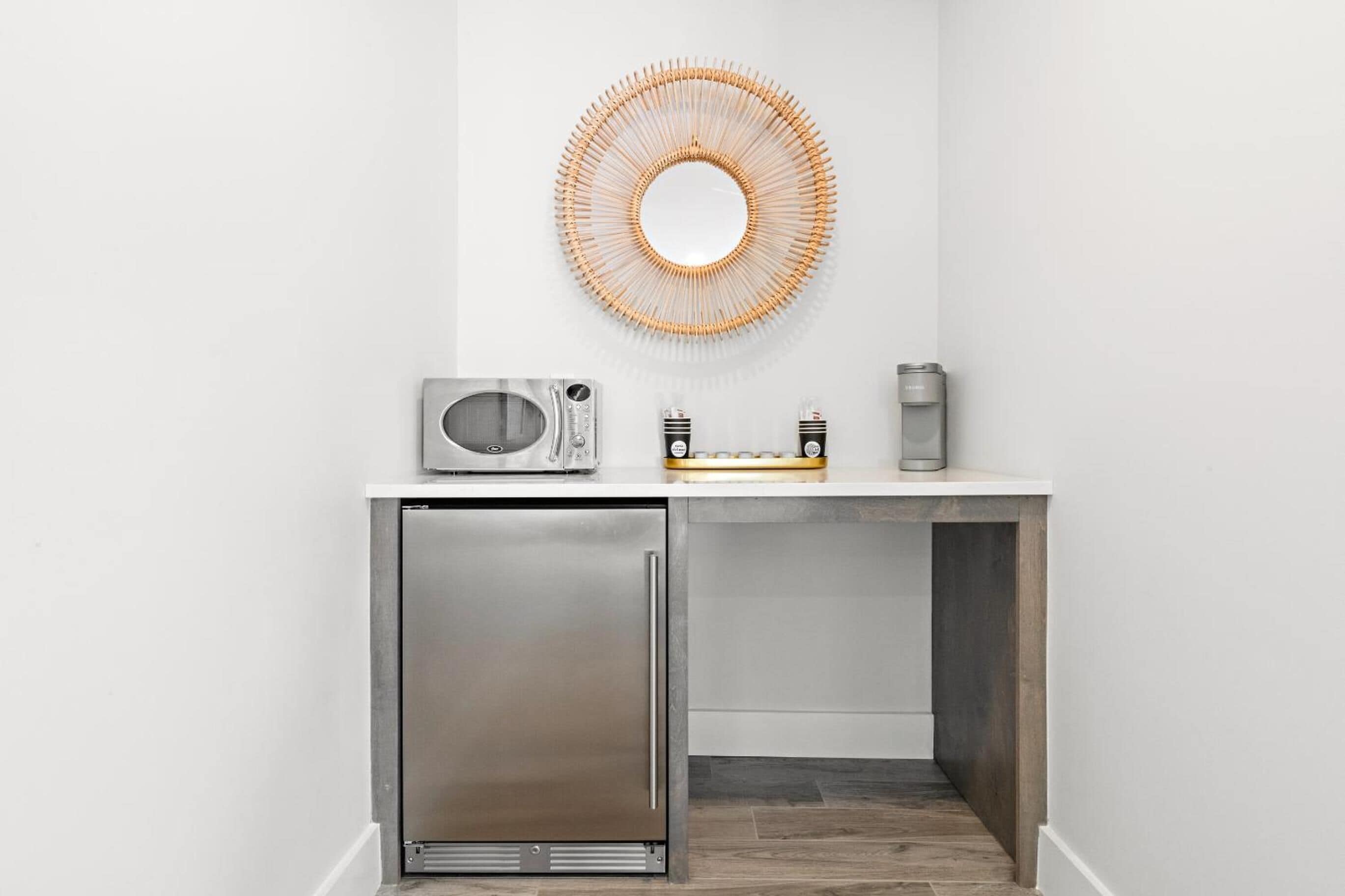 A compact kitchenette with a microwave, toaster, and coffee maker on a white countertop, a small refrigerator below, and a decorative circular wall art piece above.