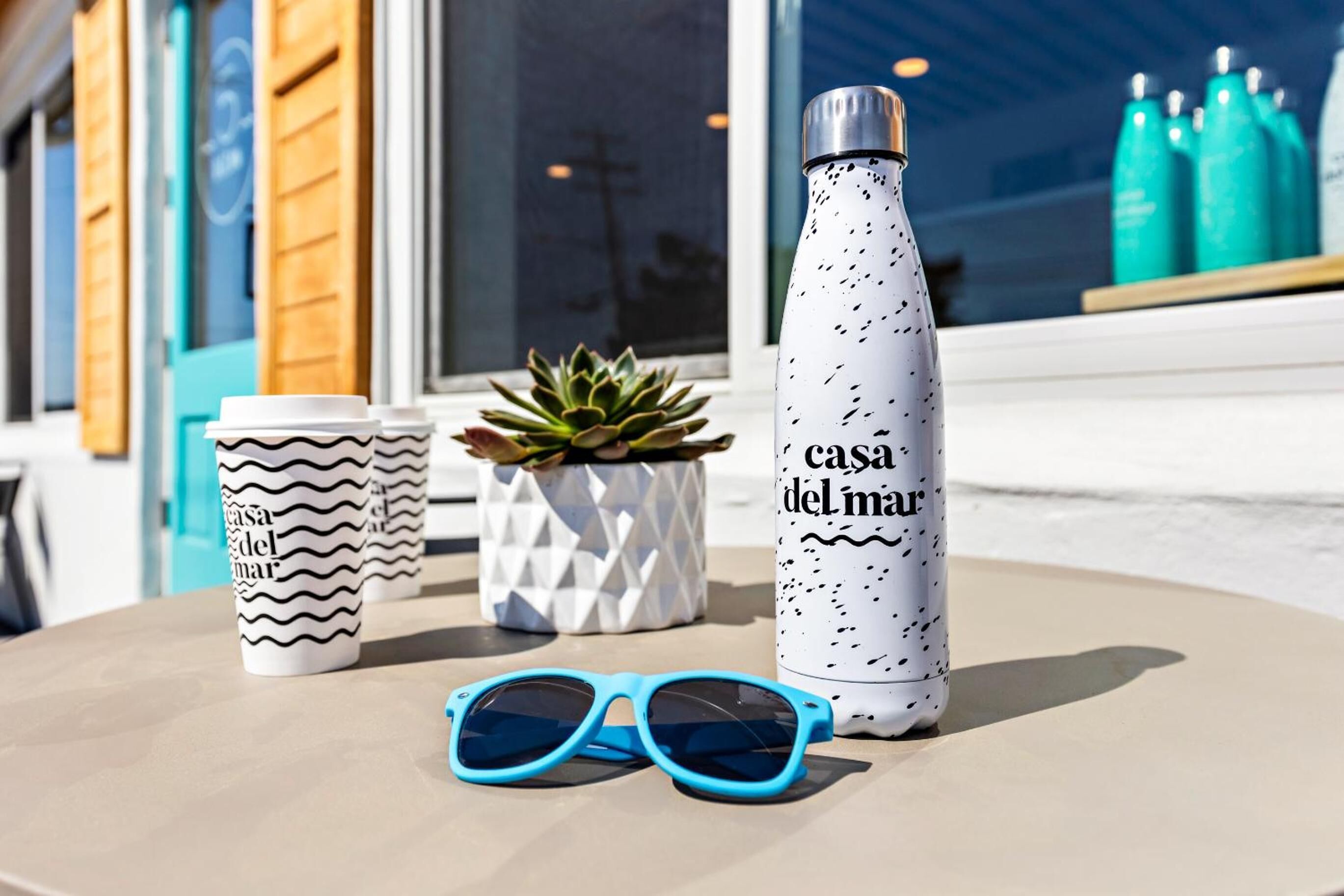 Une table avec une bouteille d’eau, deux tasses, une paire de lunettes de soleil et une petite plante.