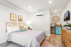 Un dormitorio con cama, mesita de noche, lámpara, equipo de aire acondicionado, televisión y una pequeña kitchenette con refrigerador.