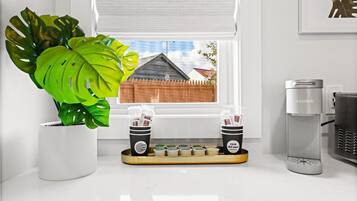 Cozinha com cafeteira, duas chávenas de café, um tabuleiro com cápsulas de café e uma grande planta em vaso.