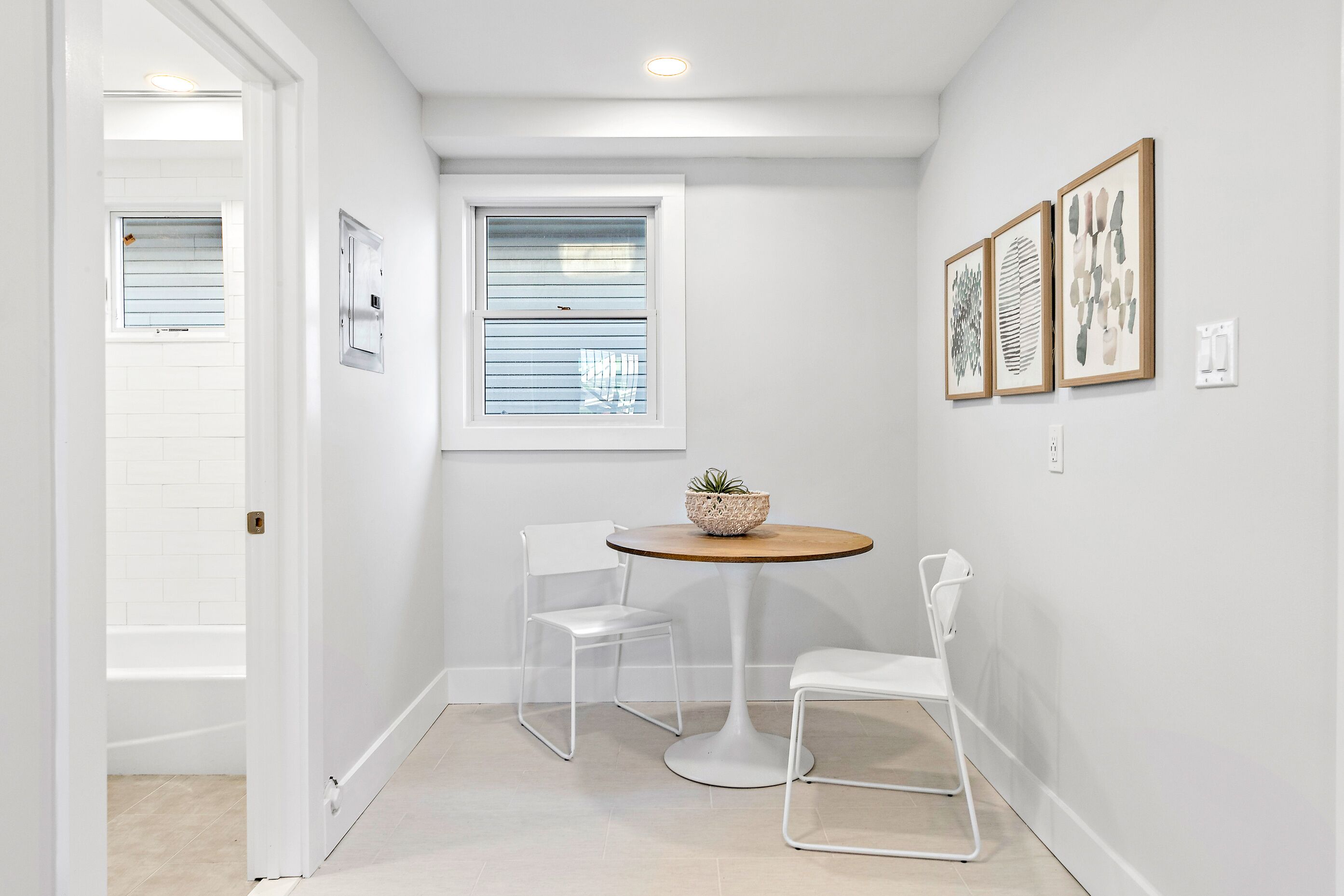 A small, well-lit room with a white door, a window with blinds, a round table with two chairs, and three framed artworks on the wall.