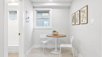 A small, well-lit room with a white door, a window with blinds, a round table with two chairs, and three framed artworks on the wall.