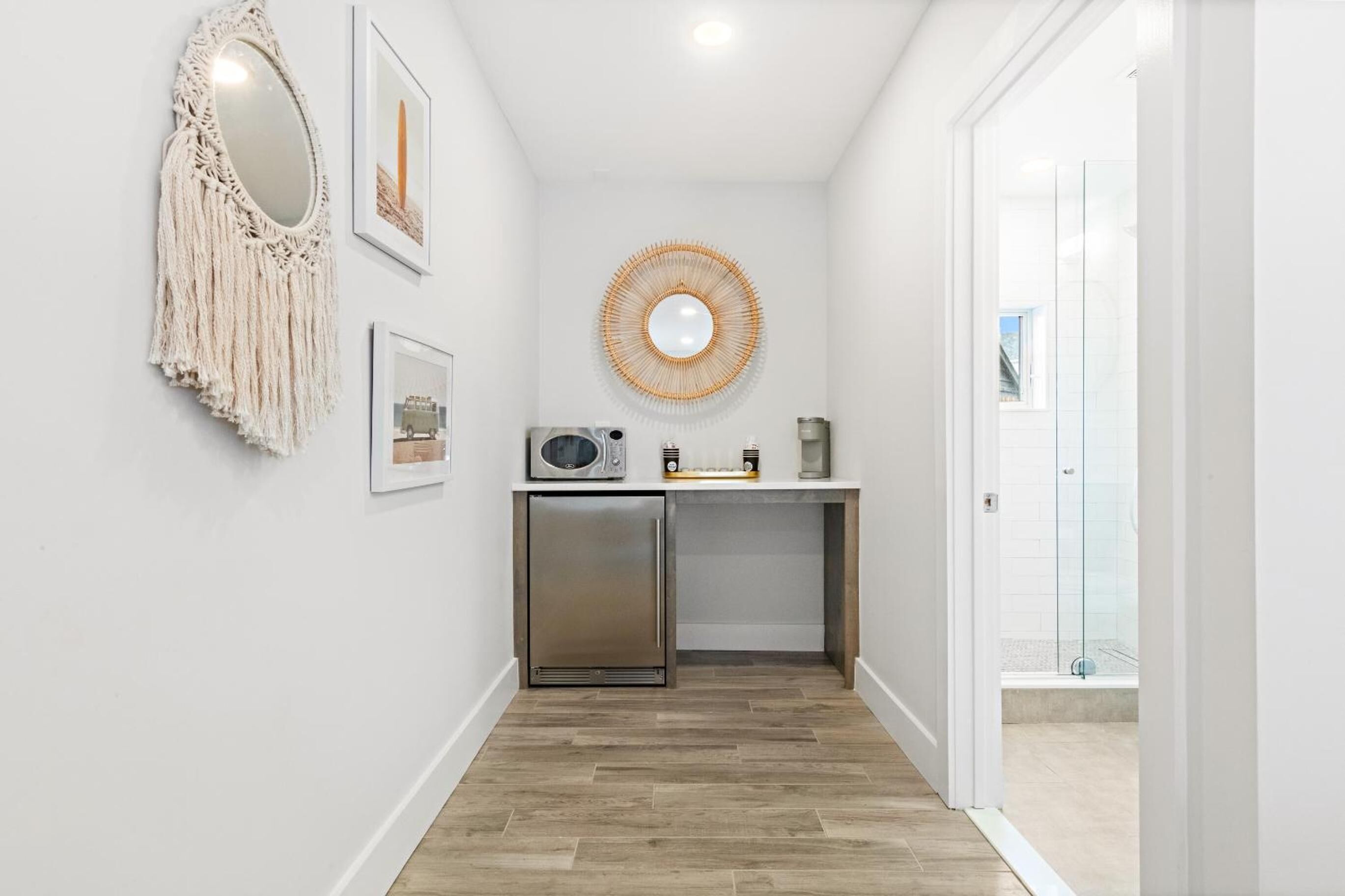 A hallway with a wooden floor, a small kitchenette with a microwave, a refrigerator, and a round mirror on the wall.