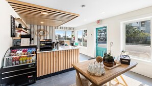 Un moderno vestíbulo de hotel con una recepción, una máquina de café, un dispensador de bebidas y vistas a la calle a través de grandes ventanales.