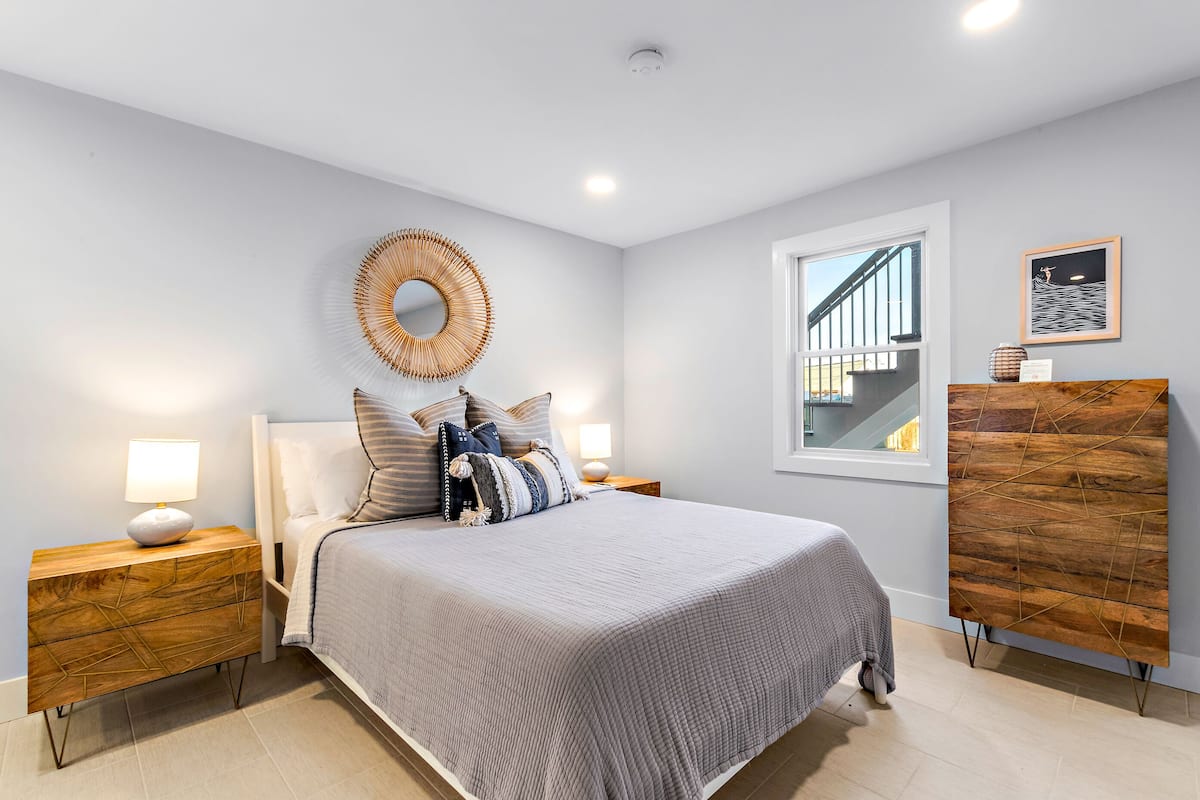 A modern bedroom with a large bed, bedside tables, a mirror, and a window with a view of a staircase.