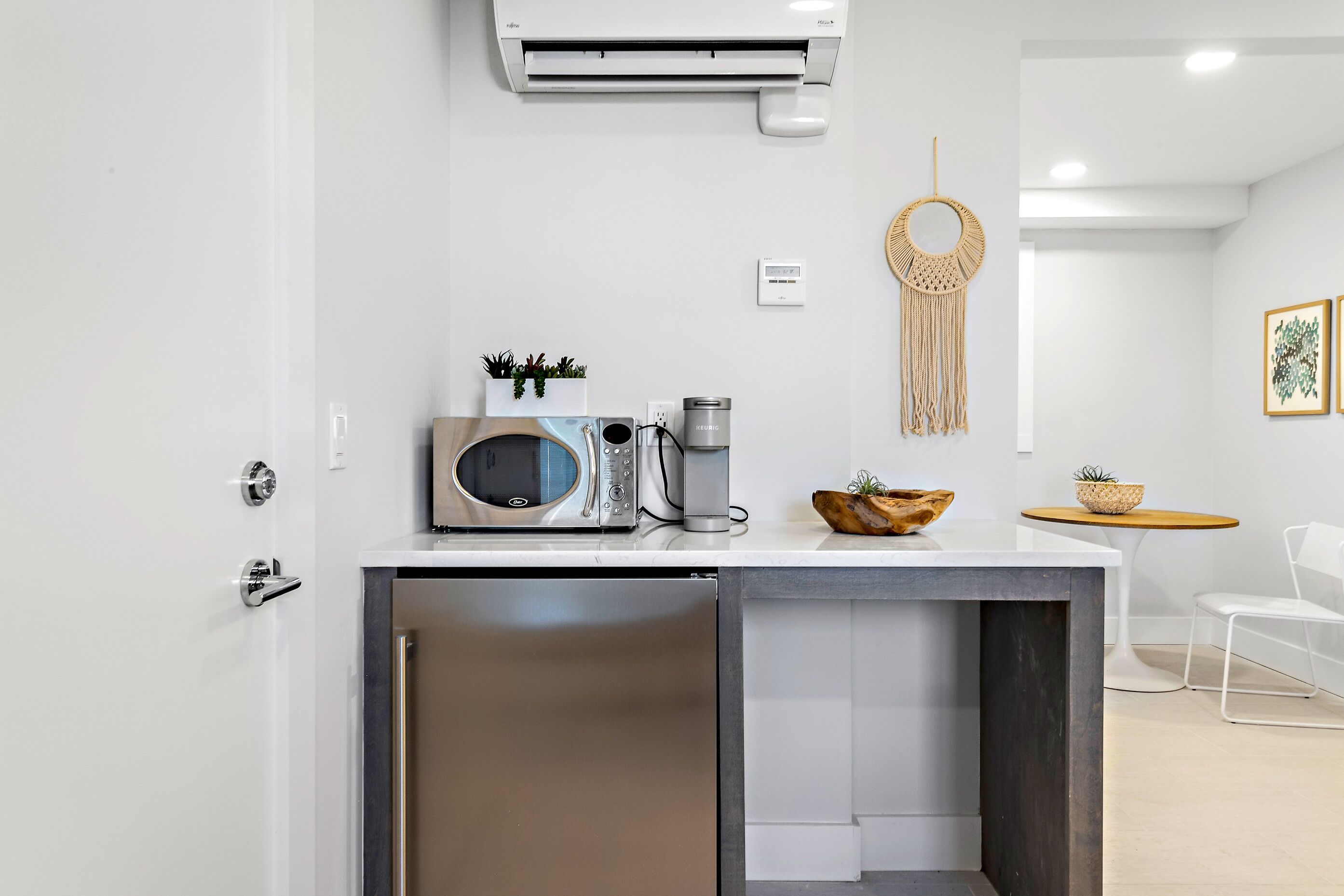 Cozinha moderna com micro-ondas embutido, máquina de café e pequena geladeira. Apresenta um pendente de macramê decorativo e uma pequena mesa com uma tigela e plantas.