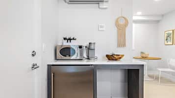 Cozinha moderna com micro-ondas embutido, máquina de café e pequena geladeira. Apresenta um pendente de macramê decorativo e uma pequena mesa com uma tigela e plantas.