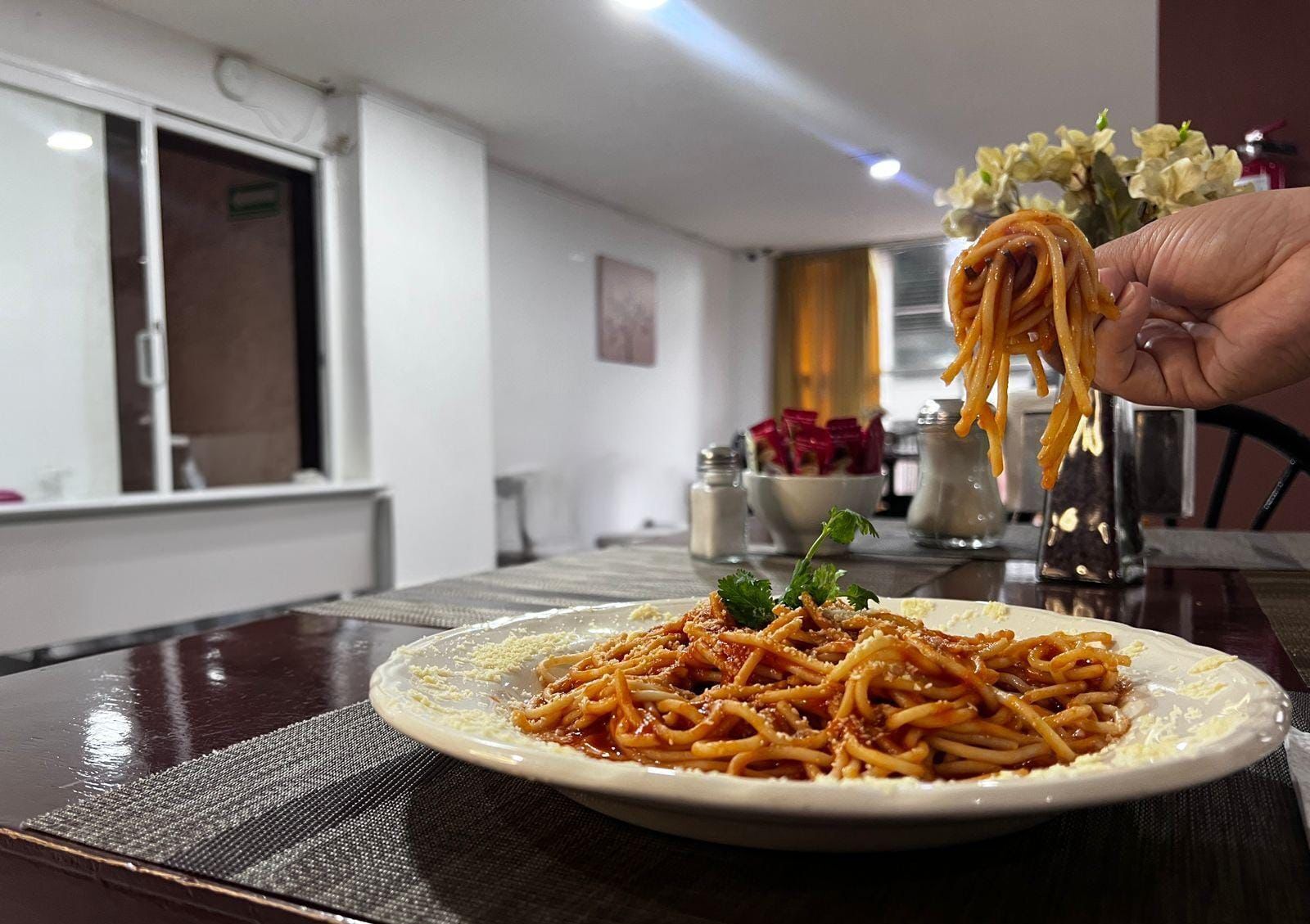 Ein Esstisch mit Spaghetti, eine Hand, die Spaghetti hält, und eine Vase mit Blumen im Hintergrund.