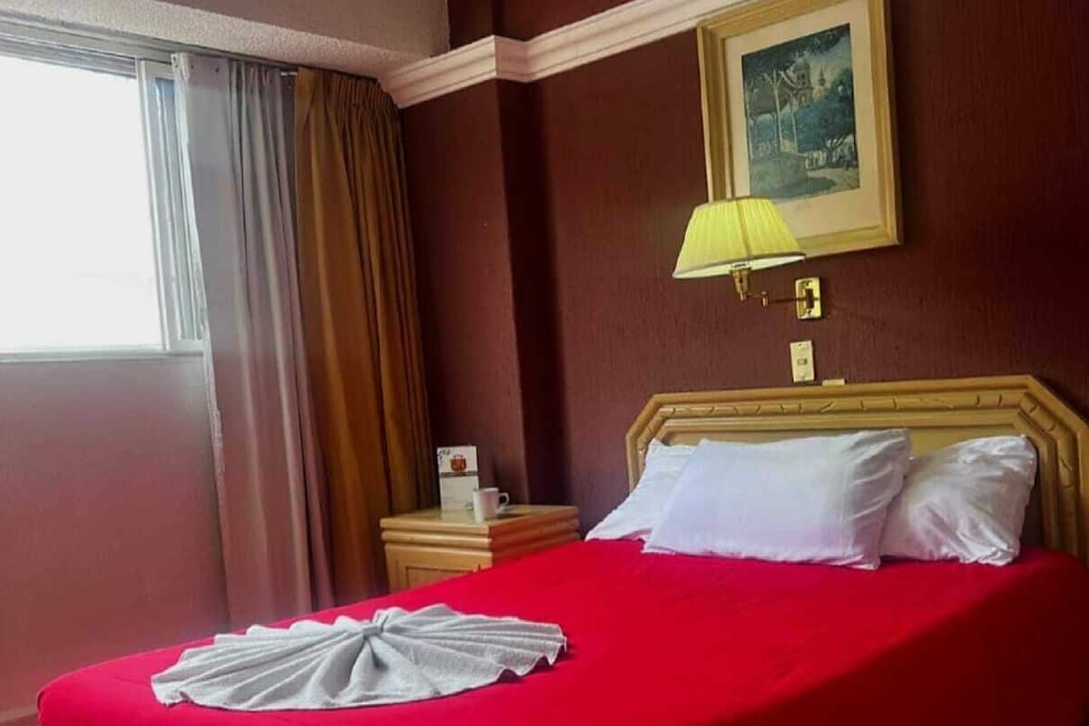 Een hotelkamer met een bed, een nachtkastje, een lamp, een raam met gordijnen en een schilderij aan de muur.
