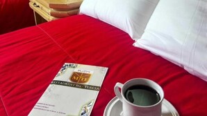 Habitación de hotel con colcha roja, almohada blanca, toalla doblada, taza de café sobre un platillo y un menú sobre la cama.